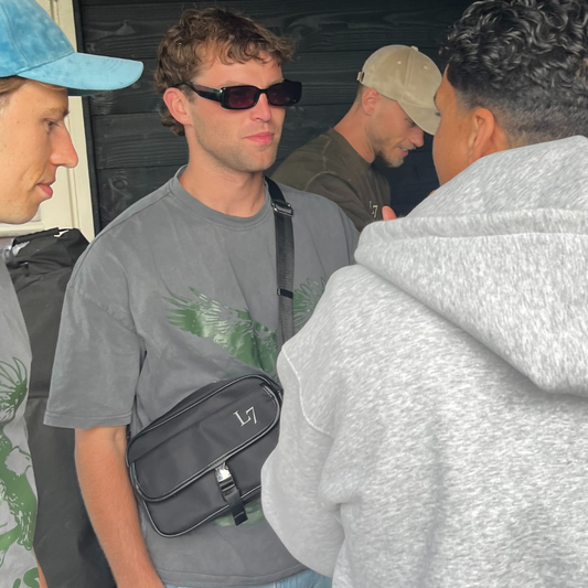 Man draagt grijs Louis Zeven boxy shirt met adelaar-print. Ook draagt hij een nylon messenger bag